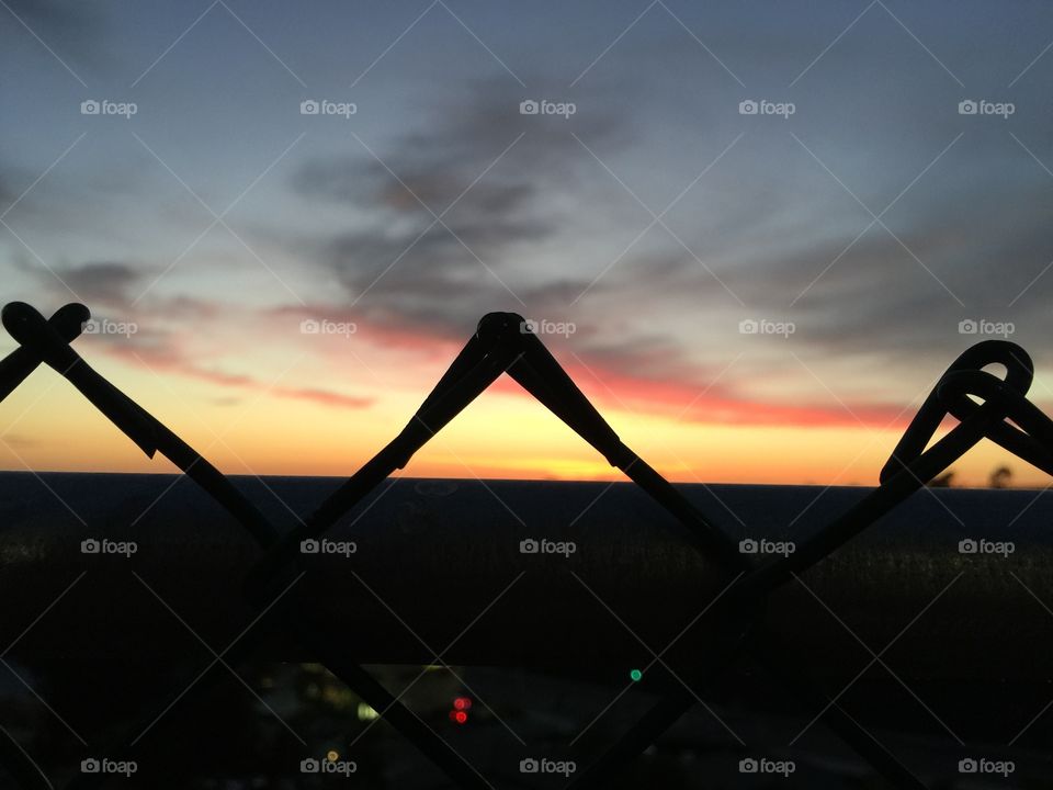 A picture taken peering over a fence into a sunset with city lights in the bottom frame