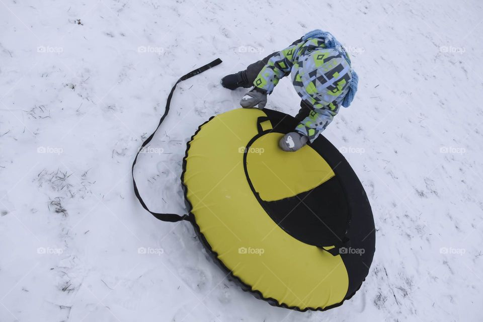 A small, carefree boy walks in the white snow in winter and rides a tubing in the park, near trees in the snow.