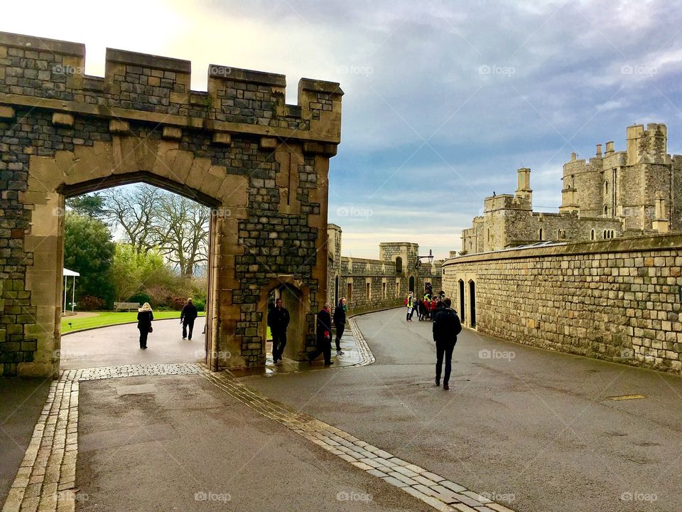 A stone entryway to Windsor Castle in England. People stroll up the hill and the sky is blue and cloudy. 