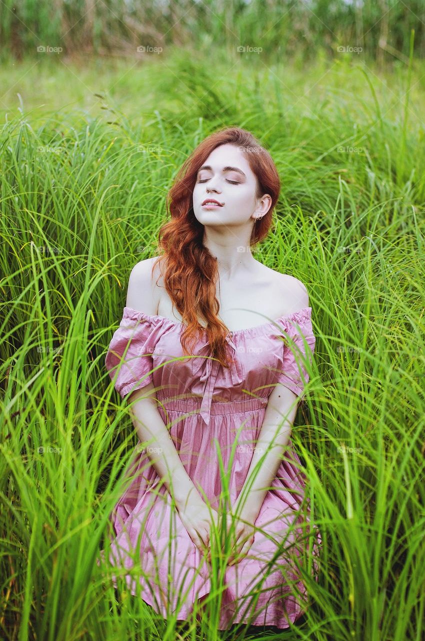 Portrait of beautiful young redhead woman, girl in pink dress sitting  outdoor, in the summer park, meadow with green grass