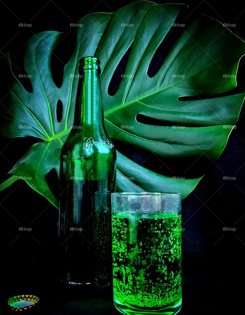 Green color. On a black background, a green leaf of Monstera, a green glass bottle and a glass with a green drink. There is a metal cork next to the bottle.
