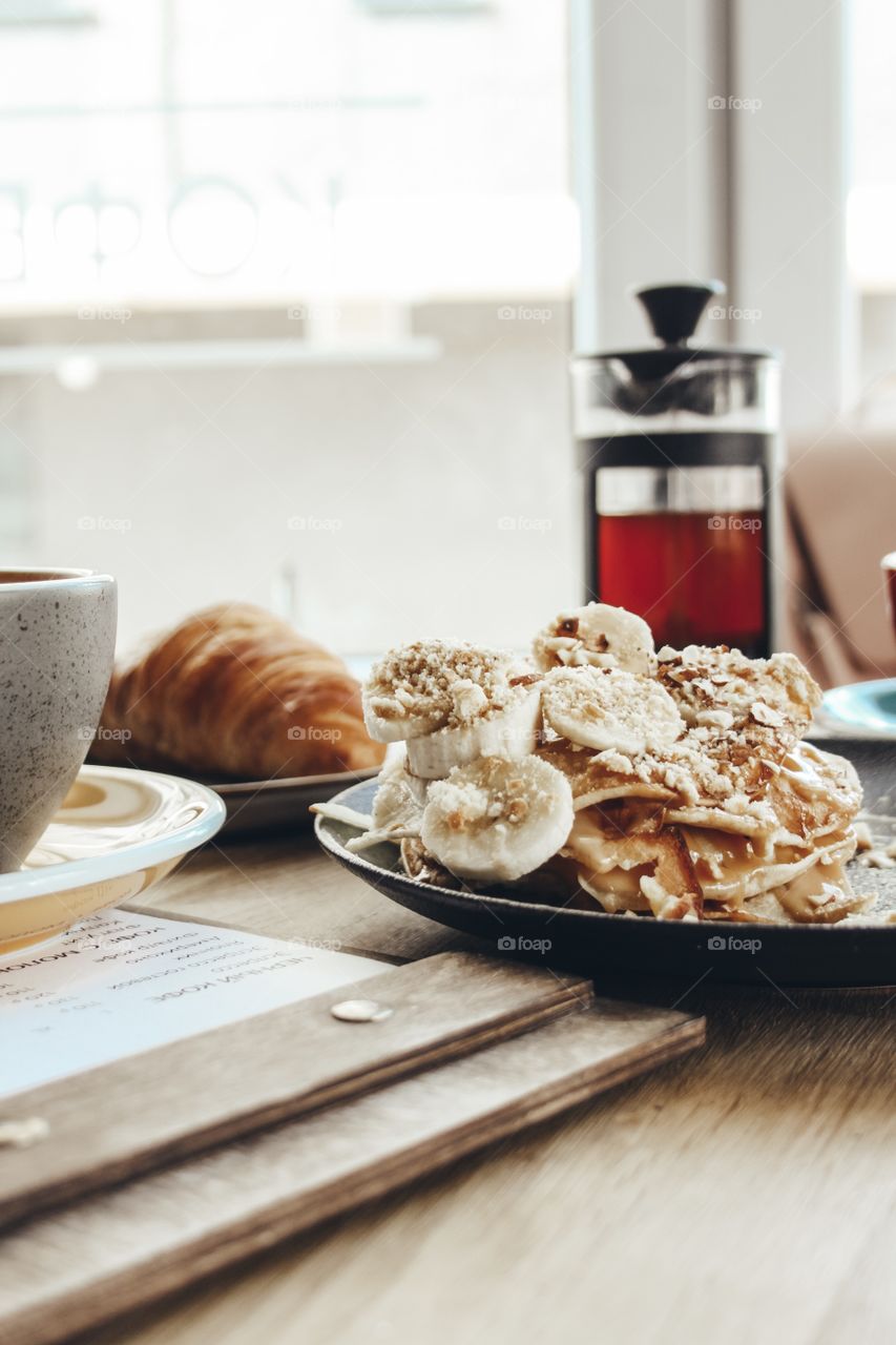 Banana pancakes, tasty croissant, cappuccino and black tea on the table in a natural light