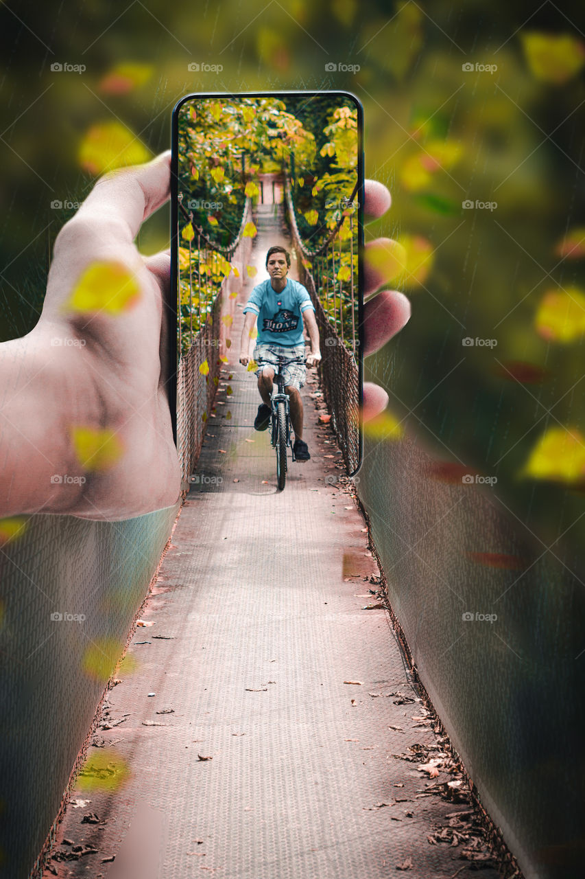 POV photography. Edition of a young man riding a bicycle on a bridge inside the forest. Attention lines of the bridge towards the subject. Subject protrudes from the phone