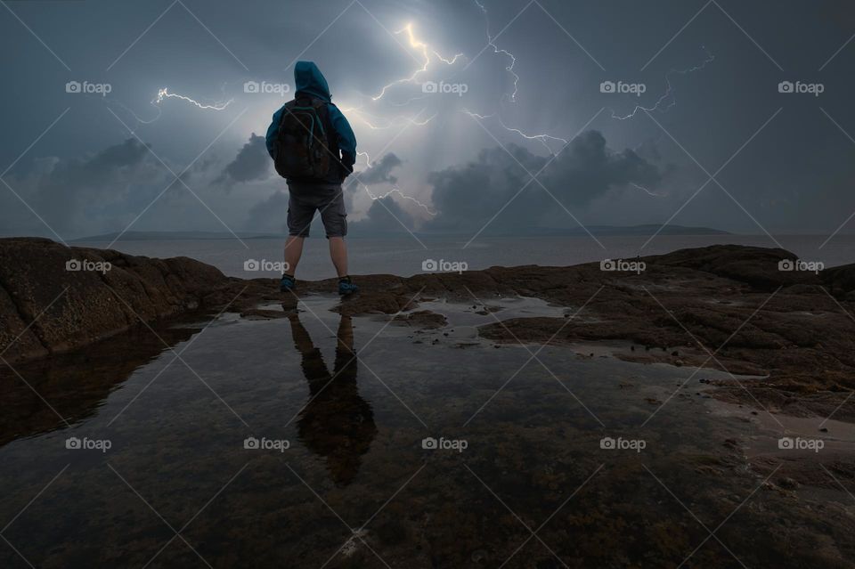 Man on the rock reflected in water looking at thunderstorm with lightning strikes over wild atlantic way in county Galway, Ireland