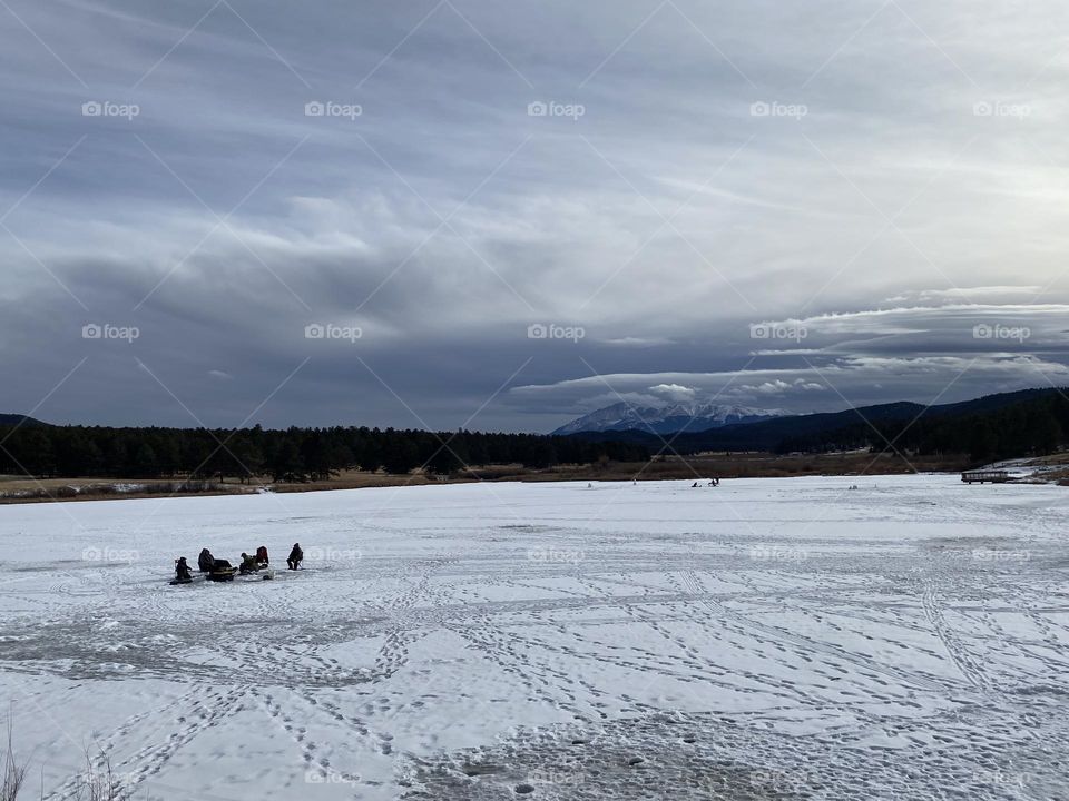 Men ice fishing on a snow covered frozen lake in the winter. Snowy Pikes peak in the background is peeking through the winter clouds.