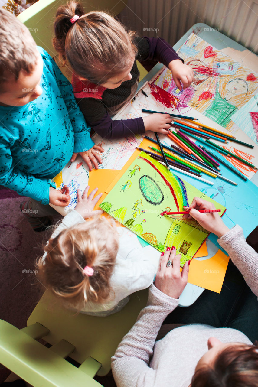 Mom with her little daughters and son together drawing a colorful picture of playing children using pencil crayons sitting at table. Photo from above