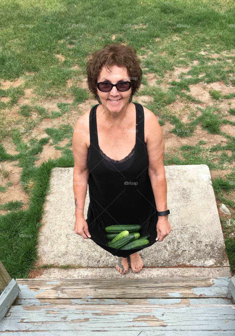 Woman standing with skirt full of cucumbers on cement landing at bottom of outside steps, smiling, sunglasses, summer, barefoot.