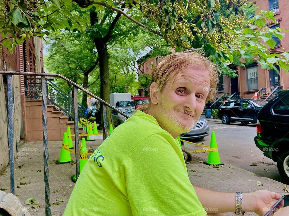 „Timothy Trespas“ my partner in life is sitting on the sidewalk of „Waverly St“ in „Clinton Hill“, Bklyn, NY. His bright neon green T-Shirt matches the color of the plastic barricades put up behind him. 2022. Hypnotic Productions