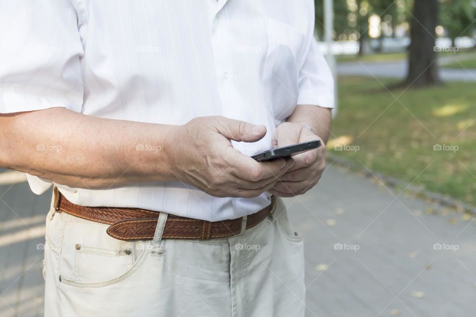 An elderly man walks alone in the park in the summer. A modern pensioner, businessman in a white shirt and trousers takes pictures with a camera in a mobile phone.