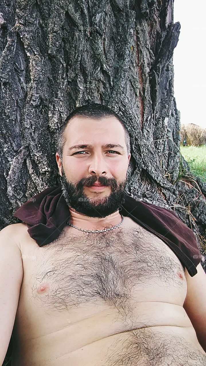 a man with a beard with a bare torso sitting under a tree