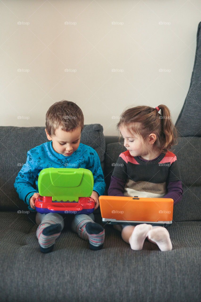 Two children playing with laptops learning basic digits, characters, sounds and images. Little girl and boy sitting on sofa and playing together at home. Candid people, real moments, authentic situations