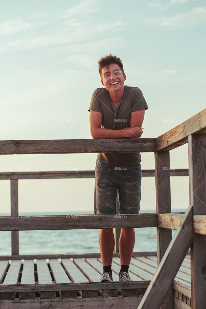 Smiling happy young man standing on a pier over the sea during summer vavations. Copy space room for text