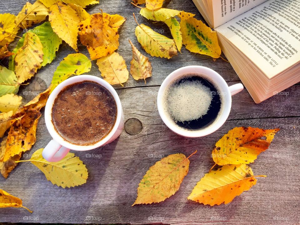 Two cups of coffee on wooden table with a book and yellow autumn leaves beside