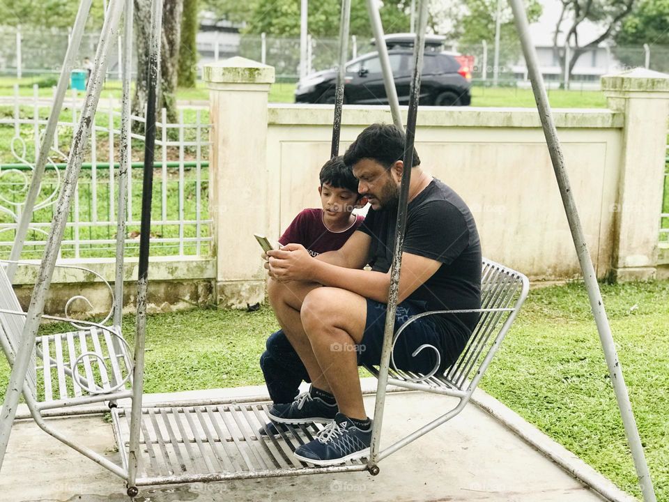 Father explaining something to son from phone by swinging outside.