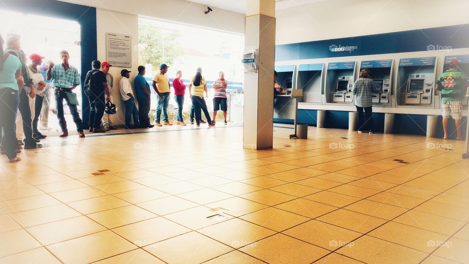 Pessoas em fila aguardando atendimento com senhas, próximas a caixas eletrônicos bancários.