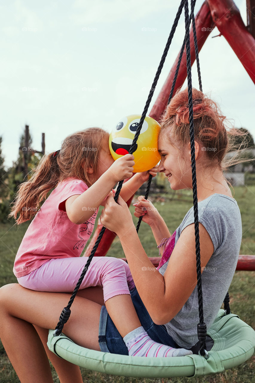 Teenage girl playing with her younger sister in a home playground in a backyard. Happy smiling sisters having fun on a swing together on summer day. Real people, authentic situations