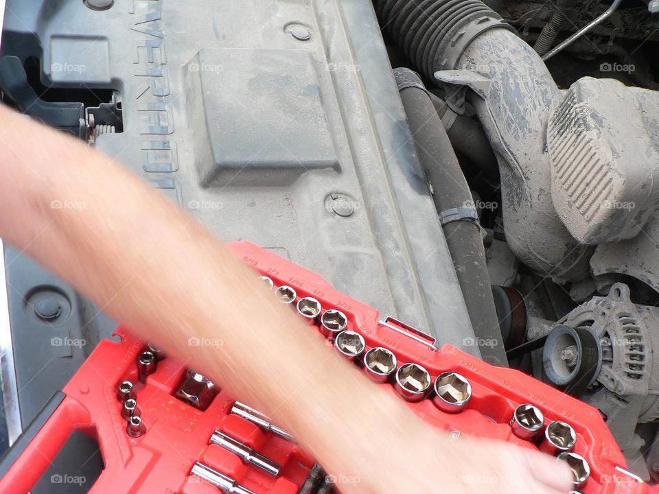 Under the hood of a red pickup truck with combination wrench set tools organized in a red tool hard case by the work station and the mechanic handyman fixing a piece.