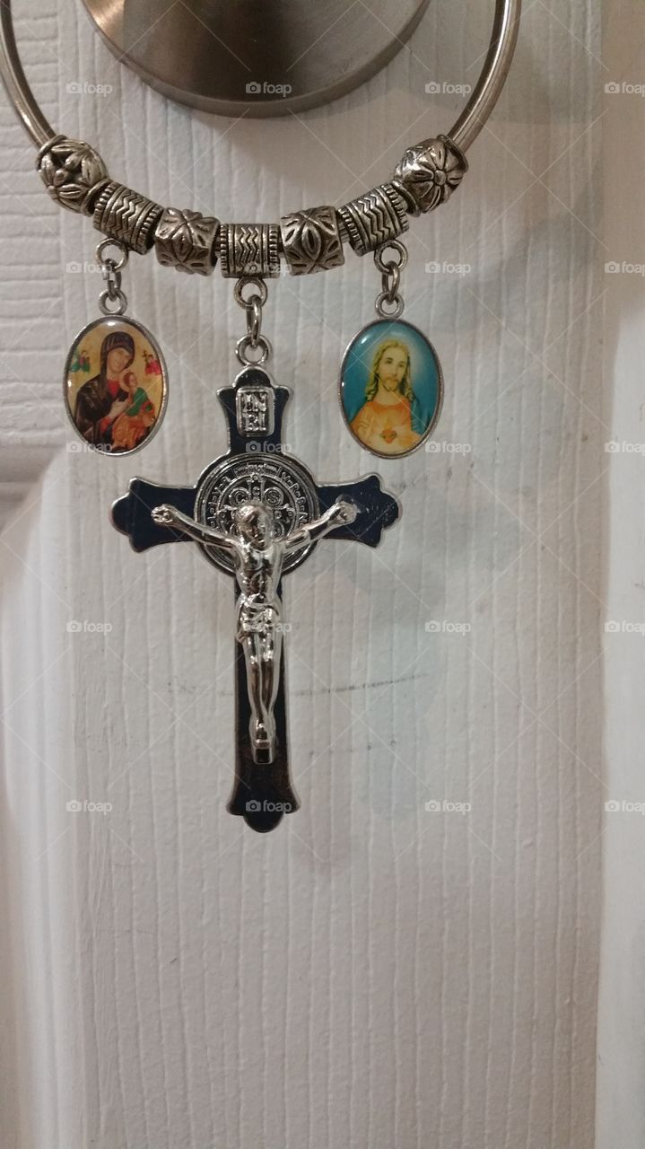close up of religious door hanger with silver cross that has st Benedicts medal in the center of the cross and 2 mdellaiona of Christmas and Madonna and child