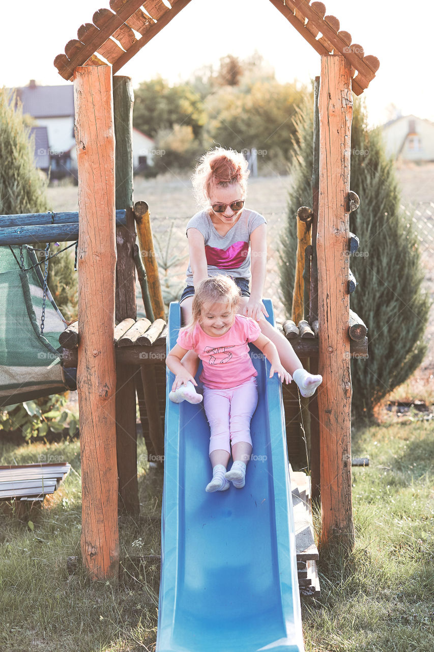 Teenage girl playing with her younger sister in a home playground in a backyard. Happy smiling sisters having fun on a slide together on summer day. Real people, authentic situations