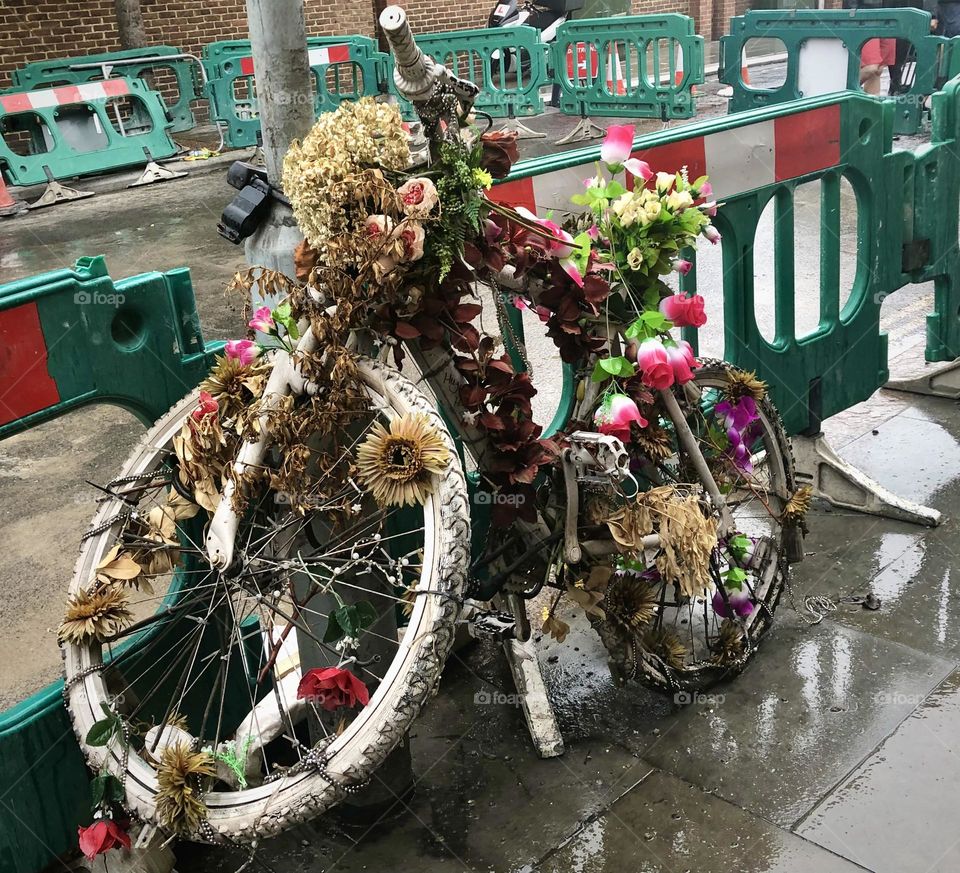 A old bicycle abandoned in the street, covered in rotting and bedraggled flowers, in front of roadworks 