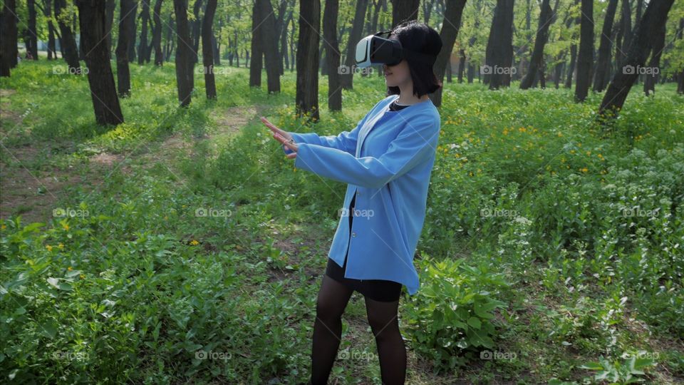 Portrait of trendy woman in blue coat using virtual reality glasses simulator in the park or forest at spring 