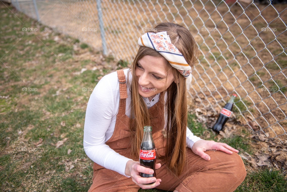 Young millennial woman sitting in the grass, laughing and holding a bottle of Coca-cola