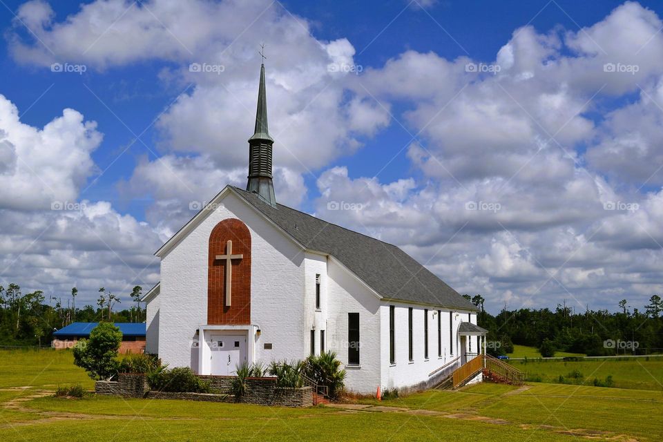 A traditional white church with a grey roof, a cross on the front, and a steeple against white puffy v cvs loads and a blue sky