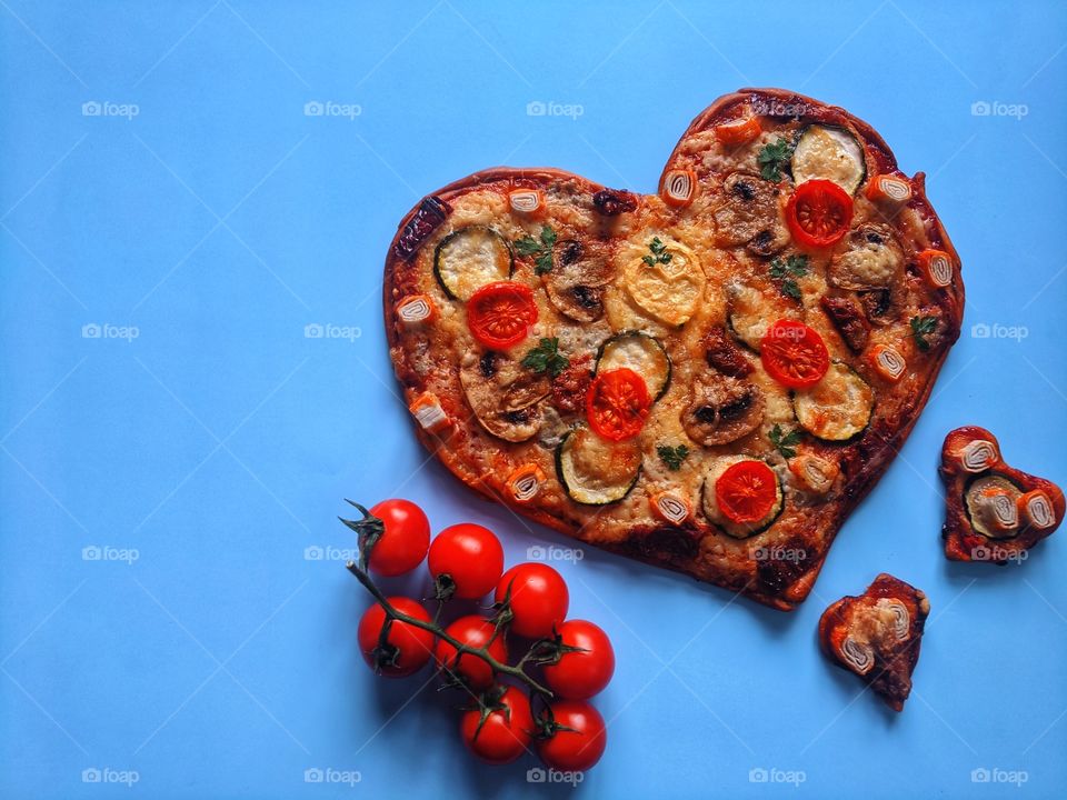 Delicious vegetarian pizza in the shape of a heart for Valentine's Day with tomatoes, vegetables and cheese on a blue background.