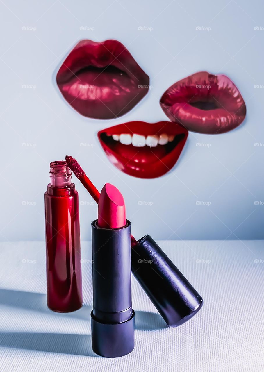 Red lipstick and lipgloss displayed in front of cut outs of red lips stuck to a white background 