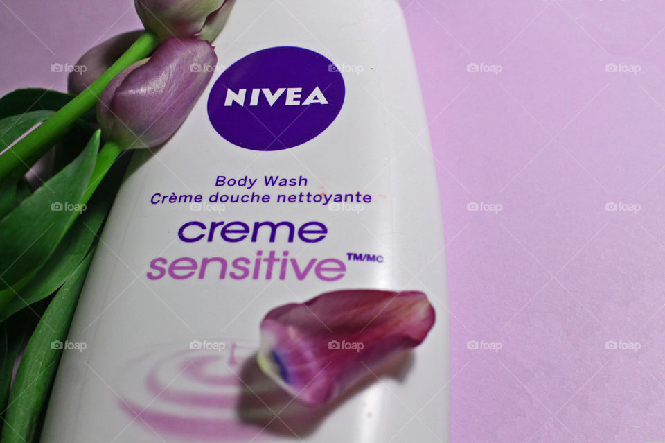 A flat-lay closeup of a bottle of NIVEA creme sensitive Body Wash with purple tulips on a pale violet background.  One delicate purple and blue tinged petal rests on the bottle. 
