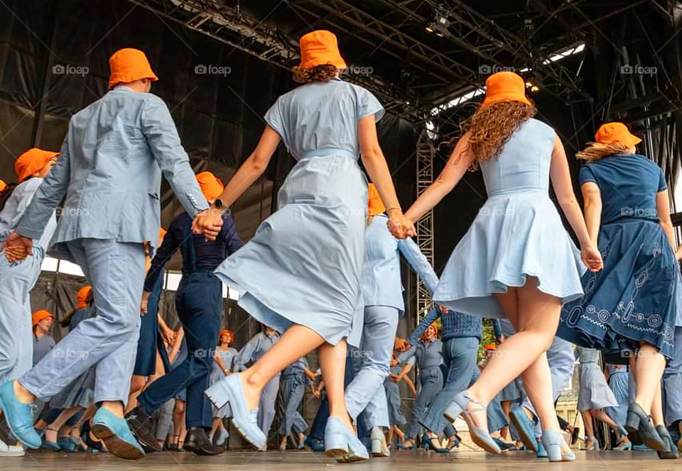 Women and men all dressed in blue with orange hats dancing hand in hand a Breton traditional dance on stage at Saint-Loup Festival in Guingamp