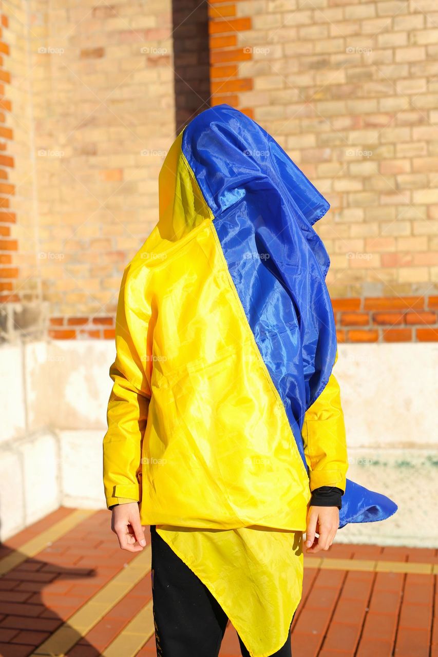 a teenager in the city center, stands with a yellow-blue flag, the child is wrapped in a flag