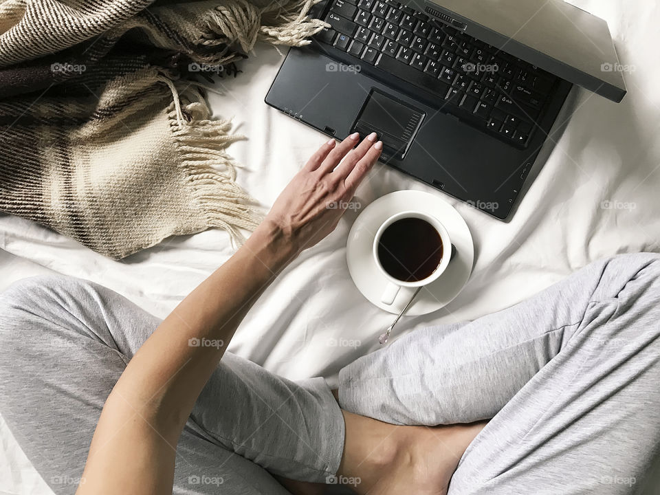 Overheard view of a young woman using her notebook and checking social media with a cup of coffee in cozy bed 