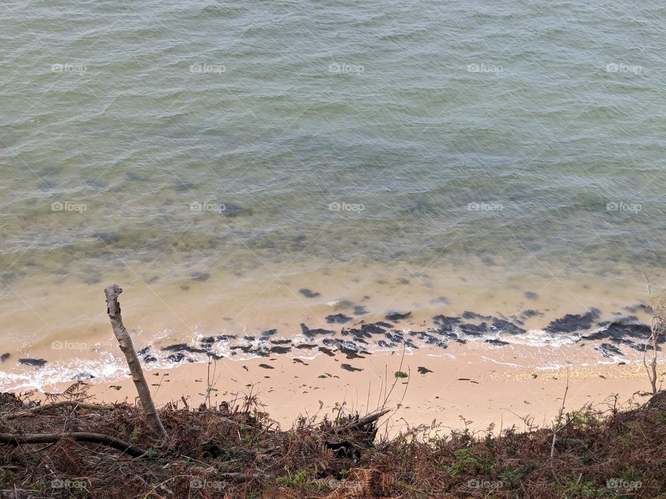 Looking down from a hill at clear, calm water on a sunny day. The sand has a line of rocks at the waters' edge