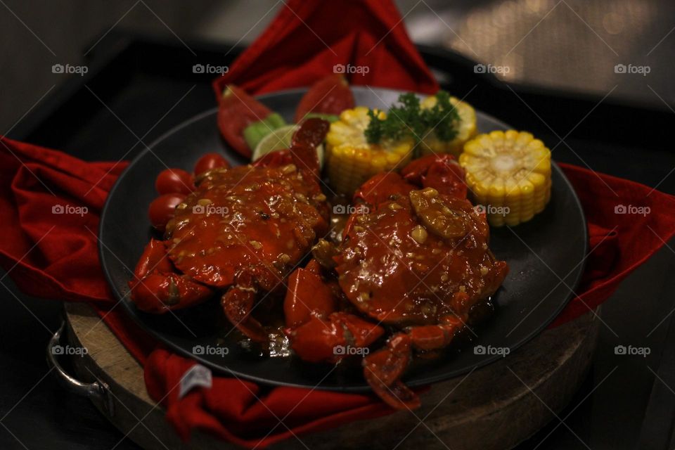 Crab flavored in creamy garlic sauce served with corn, tomatoes, lemon wedges and cherries on a black plate set on a red cloth set in a wooden setting. Asian flavor crab.