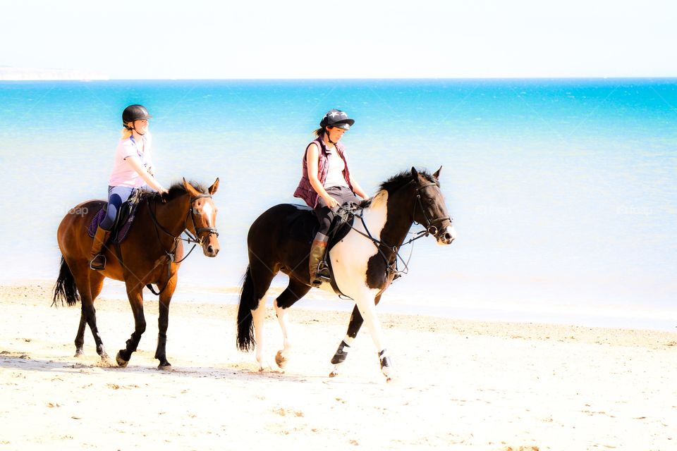 A pair of horses or ponies with their riders walking along a white sandy beach with the ocean behind.