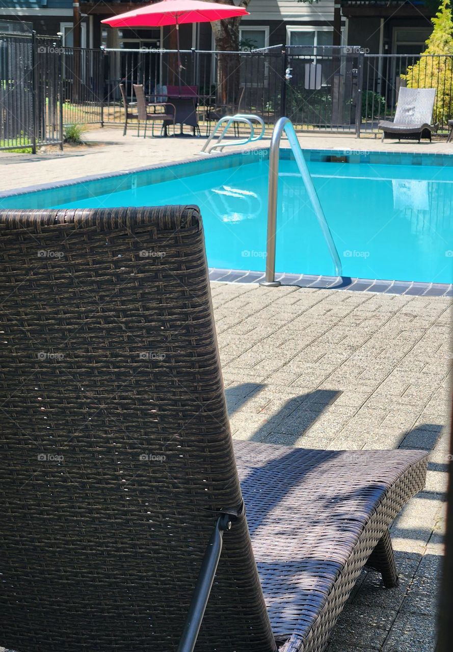 view from a wicker lounge chair in the shade of a bright blue swimming pool and cozy umbrella covered table in the corner on a sunny Summer day