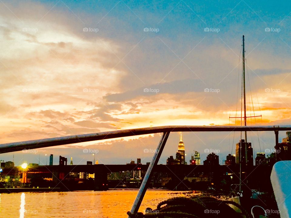 A view of the Manhattan skyline featuring the illuminated Empire State Building in the far distance beyond the waters of the East River at Newtown Creek that reflect at this hour the golden rays of the setting sun. Photo 2018. Hypnotic Productions