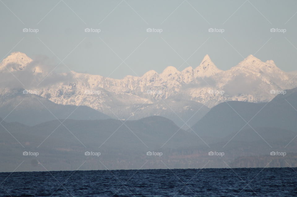 Shot of the Northwest Pacific Coast glowing with the rare winter sunlight. The sea is indigo blue, the hills behind dark green,  mountains capped with fresh snow and small wisps of powdery clouds kiss the mountain peaks. 