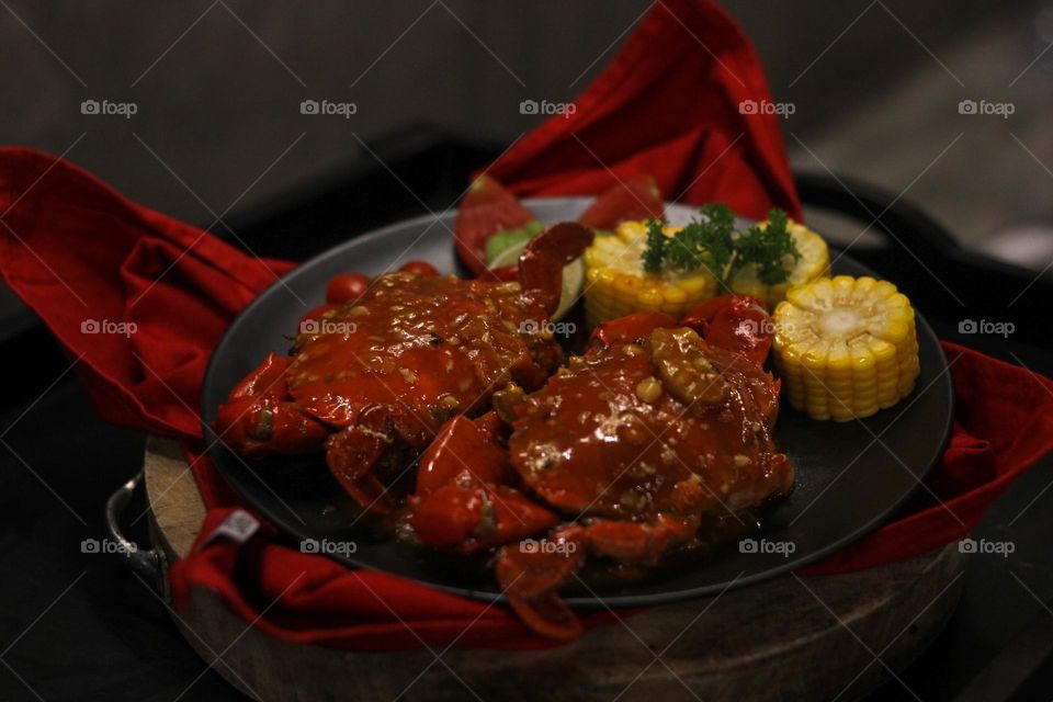 Crab flavored in creamy garlic sauce served with corn, tomatoes, lemon wedges and cherries on a black plate set on a red cloth set in a wooden setting. Asian flavor crab.