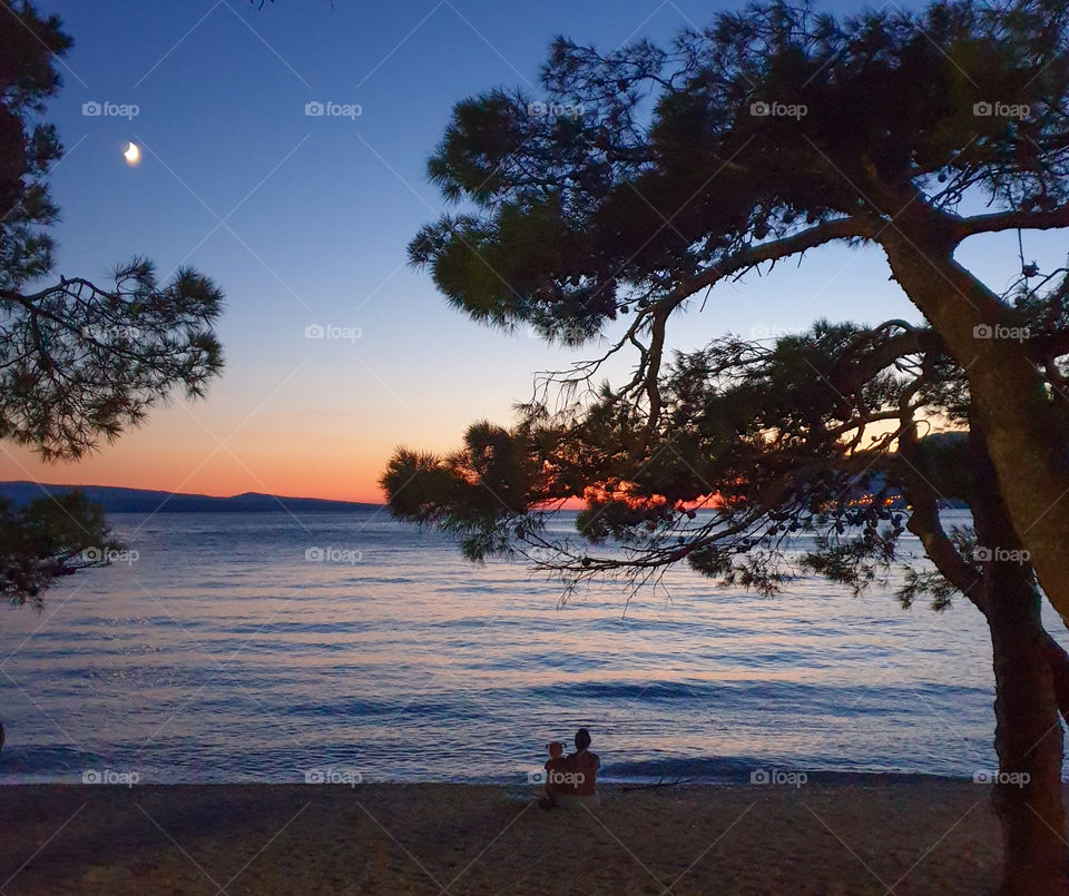 Croatia, Dalmatia, Brela, Punta Rata beach, mother and little daughter sit in the evening on the seashore and watch a beautiful sunset.  Seascape with silhouettes and sunset