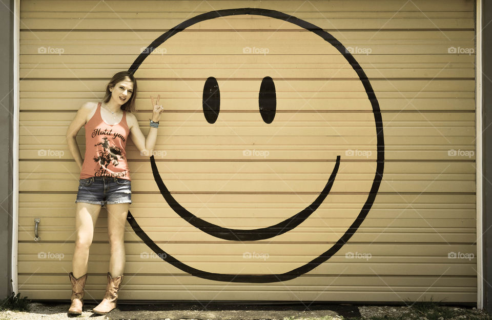 Woman standing in front of a large smiley face mural while giving a peace sign