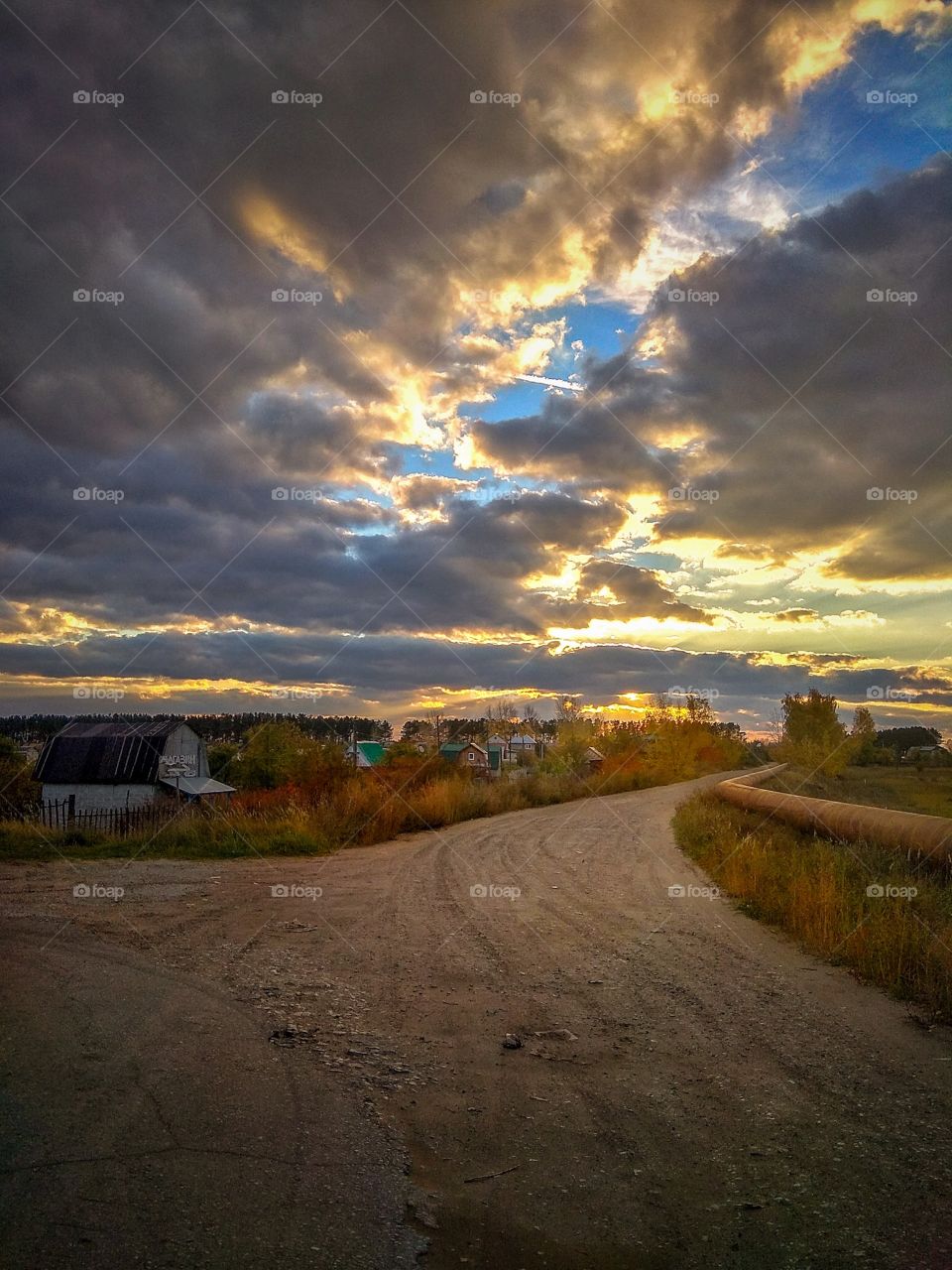 Beautiful view of the road leading to the dacha village and the houses of this village under the magnificent evening sky covered with clouds and breaking through the clouds by the rays of the setting sun in the autumn evening