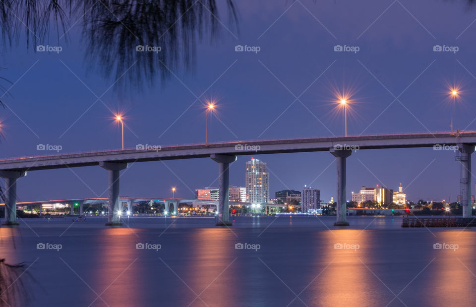bridge over water with bridge lights reflected on the water and the city buildings in the background at blue hour