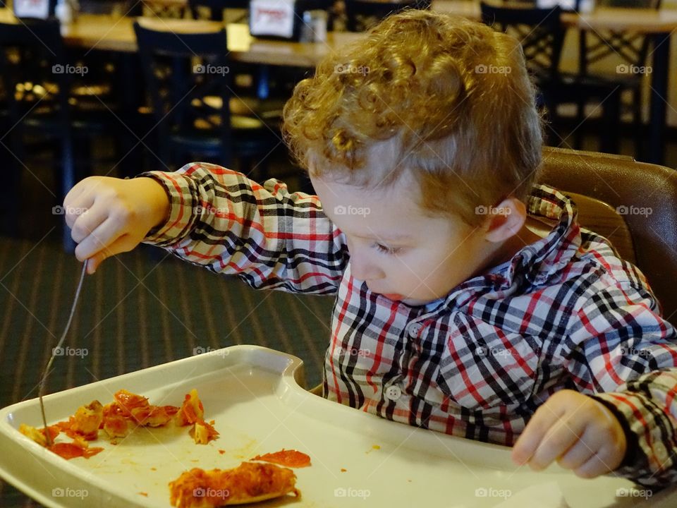 A little boy sitting in a high chair digs into his favorite snack of pepperoni pizza with great vigor. 