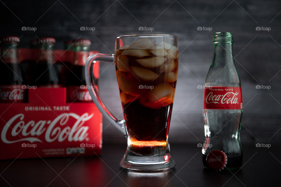 Backlit tall glass of coca-cola over ice next to an empty glass bottle of Coca-cola and a six-pack