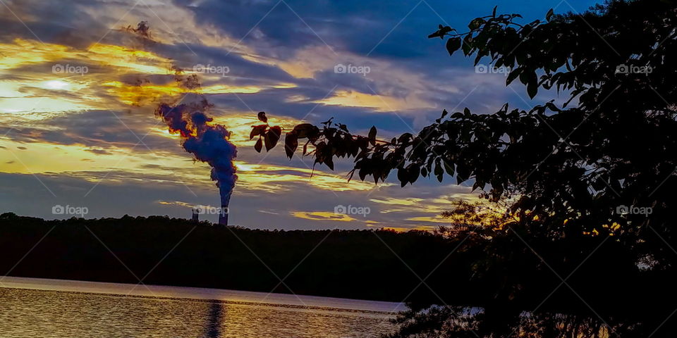 Sunset vibrant colors with power plant steam station in distance and silhouette of trees in front, all looking over water with reflection.