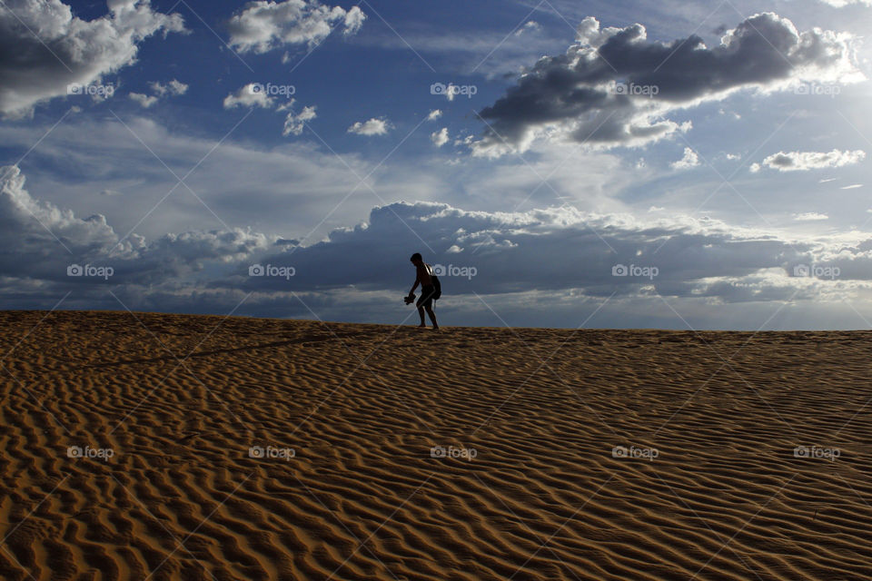 silhouette of a boy walking through a big bank of sand in contrast with a beautiful blue sky in a sunny day