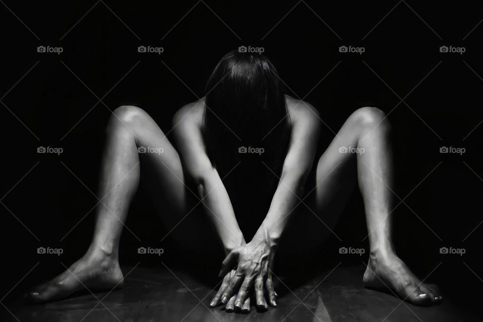 Black and white view of nude woman sitting on ground with hair, hands and legs in front.