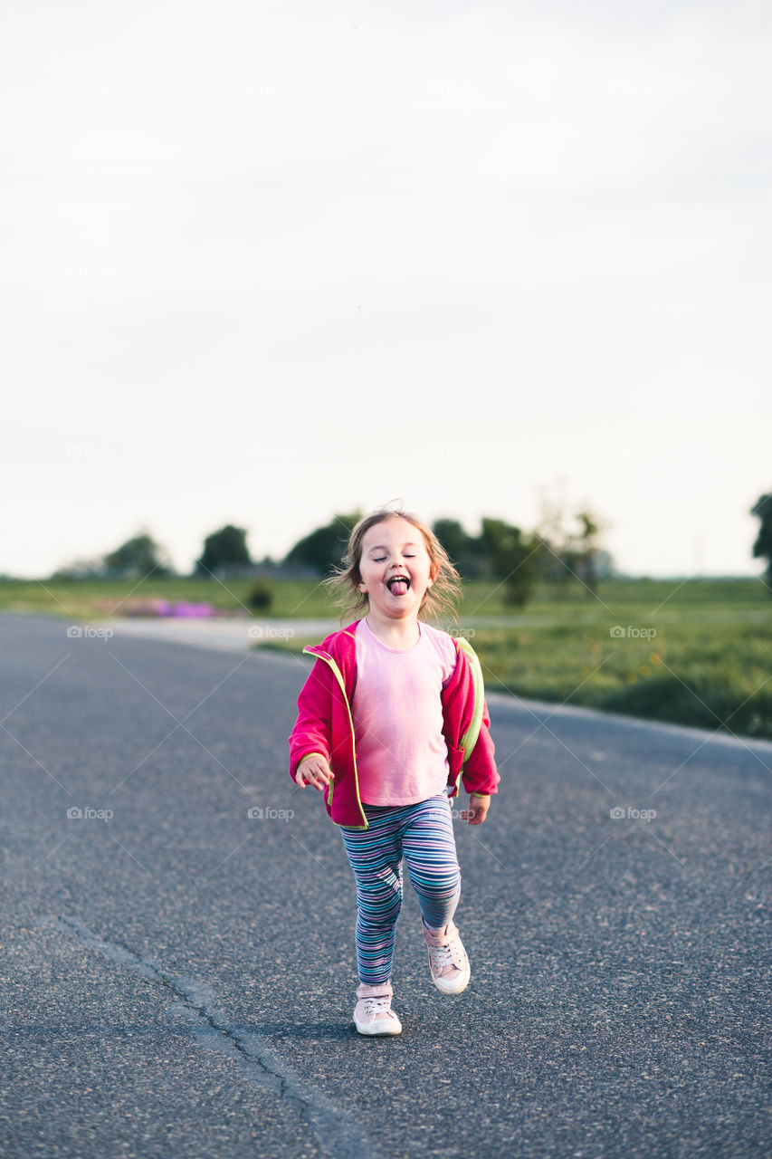 Little adorable girl having fun running on road, sticking her tongue out, playing outdoors